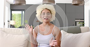 Young biracial woman with curly blonde hair gestures while speaking at home on video call on couch