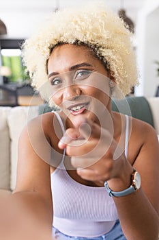 Young biracial woman with curly blonde hair is pointing and smiling at the camera on a video call