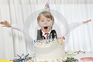 Young birthday wearing party hat with arms outstretched blowing candles on cake