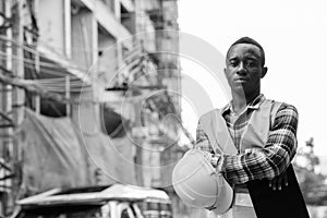 Young black African man construction worker holding clipboard and hard hat with arms crossed at building site