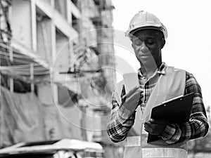Young black African man construction worker holding clipboard while using mobile phone at building site