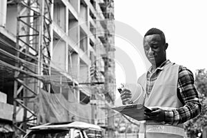 Young black African man construction worker reading on clipboard while holding hard hat at building site