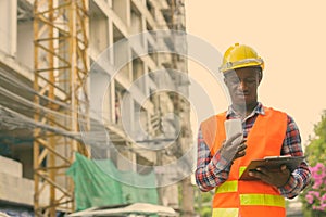 Young black African man construction worker reading on clipboard while holding mobile phone at building site