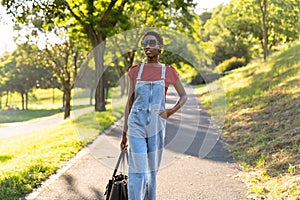 Young black African woman fashion model walking in a park with sunglasses and a handbag.