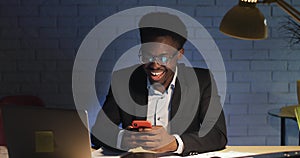 Young black businessman laughing cheerful reading funny text messages using smartphone at night office.