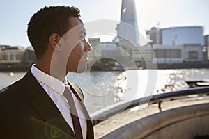 Young black businessman wearing shirt and tie standing by the River Thames, London, looking away, backlit