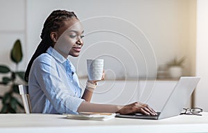 Young black businesswoman drinking morning coffee while working on laptop in office
