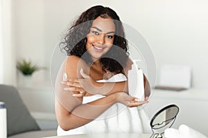 Young black chubby lady applying moisturizing body lotion on shoulder, using nourishing cream for skin