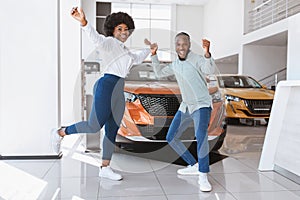 Young black couple cheering and dancing, gesturing YES, celebrating purchase of new car at auto dealership