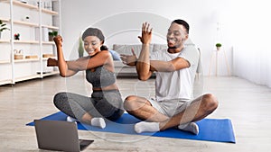 Young black couple practicing yoga or pilates, sitting in lotus pose, stretching their arms in front of laptop at home