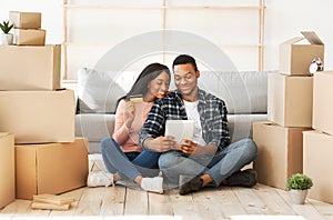 Young black couple using laptop computer and credit card for online shopping at their new home on moving day