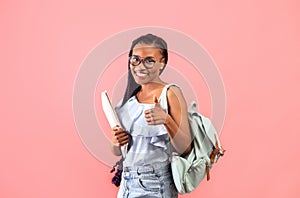 Young black female student with books and backpack showing thumb up gesture, smiling at camera over pink background