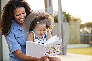 Young black girl reading book sitting on mumï¿½s knee outdoors