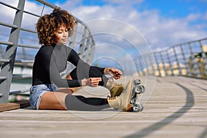 Young black girl sitting on urban bridge and puts on skates. Woman with afro hairstyle rollerblading on sunny day