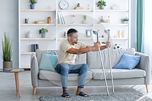 Young black guy having difficulty standing up from sofa, leaning on crutches at home