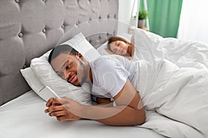 Young black guy lying on bed and using mobile phone to text his lover while his wife is asleep. Relationship infidelity