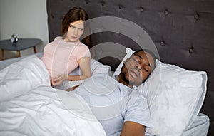 Young black guy snoring and bothering his angry sleepless wife in bed at home