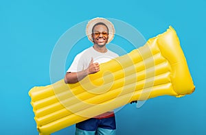 Young black guy in summer wear and straw hat holding yellow inflatable lilo and showing thumb up on blue background