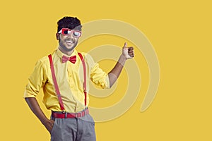 Young black haired Indian man in funny glasses for party shows thumb up stands on yellow background