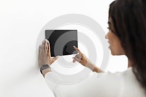 Young black lady controlling house with digital touch screen panel installed on white wall in the living room, mockup