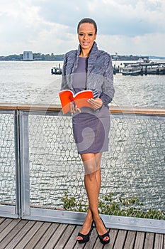 A young black lady is reading a red book on the dock by a river