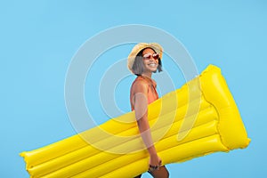 Young black lady in swimwear and straw hat holding yellow inflatable lilo on blue studio background