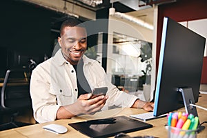 Young Black Male Advertising Marketing Or Design Creative In Modern Office Sitting At Desk Using Mobile Phone