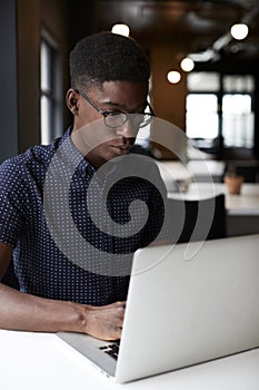 Young black male creative sitting at a desk in an open plan office using laptop, close up, vertical