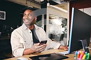 Young Black Male Smiling Advertising Marketing Or Design Creative In Modern Office Sitting At Desk Using Mobile Phone