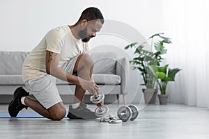 Young black man with beard in white sportswear changes weight on dumbbells in living room interior