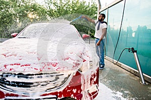 Young black man cleaning his red luxury car with foam spray outside at self car wash station. Casual African guy washing