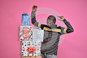 young black man feeling excited and happy and delighted, celebrating while standing next to a stack of gift boxes with fists in photo