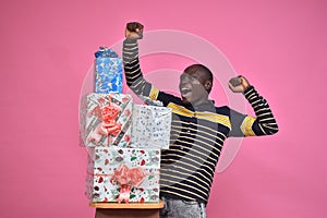young black man feeling excited and happy and delighted, celebrating while standing next to a stack of gift boxes with fists in photo