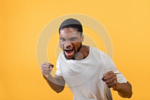 Young black man with fists defense gesture, shouting with rage, clenching fists on yellow studio background