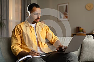 Young black man in headphones having online business conference or webinar on laptop, taking notes at home