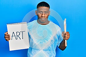 Young black man holding art notebook and painter brushes relaxed with serious expression on face