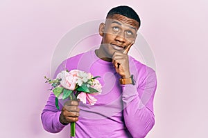 Young black man holding bouquet of white flowers serious face thinking about question with hand on chin, thoughtful about