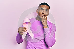 Young black man holding pink cancer ribbon serious face thinking about question with hand on chin, thoughtful about confusing idea