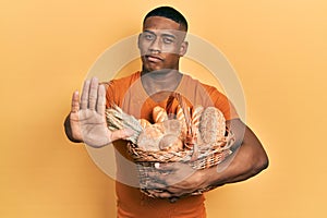 Young black man holding wicker basket with bread with open hand doing stop sign with serious and confident expression, defense