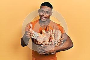 Young black man holding wicker basket with bread smiling happy and positive, thumb up doing excellent and approval sign