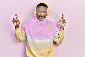 Young black man listening to music wearing headphones gesturing finger crossed smiling with hope and eyes closed