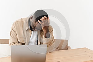 Young black man overworked, suffering from headache while sitting at laptop at table at home.