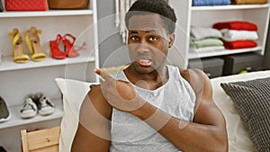 A young black man pointing while sitting in a bedroom with shelves of shoes and neat beddings photo