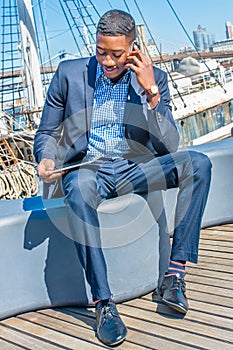 Young black man sitting by river in New York City, reading tablet computer, talking on cell phone