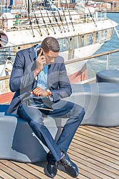 Young black man sitting by river in New York City, reading, talking on cell phone, looking at wristwatch