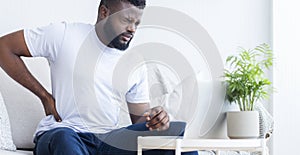 Young black man touching his sore back, working on laptop
