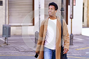 Young black man walking down the street carrying a briefcase and a smartphone in his hand.