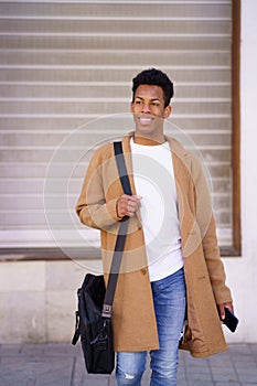 Young black man walking down the street carrying a briefcase and a smartphone in his hand.