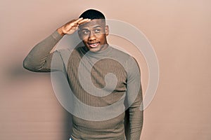 Young black man wearing casual turtleneck sweater very happy and smiling looking far away with hand over head