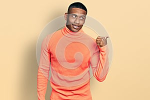 Young black man wearing orange turtleneck sweater smiling with happy face looking and pointing to the side with thumb up